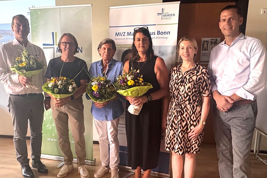 Sechs Personen stehen nebeneinander in einem Raum, drei von ihnen halten Blumensträuße, im Hintergrund zwei aufgestellte Banner mit Text und Logo